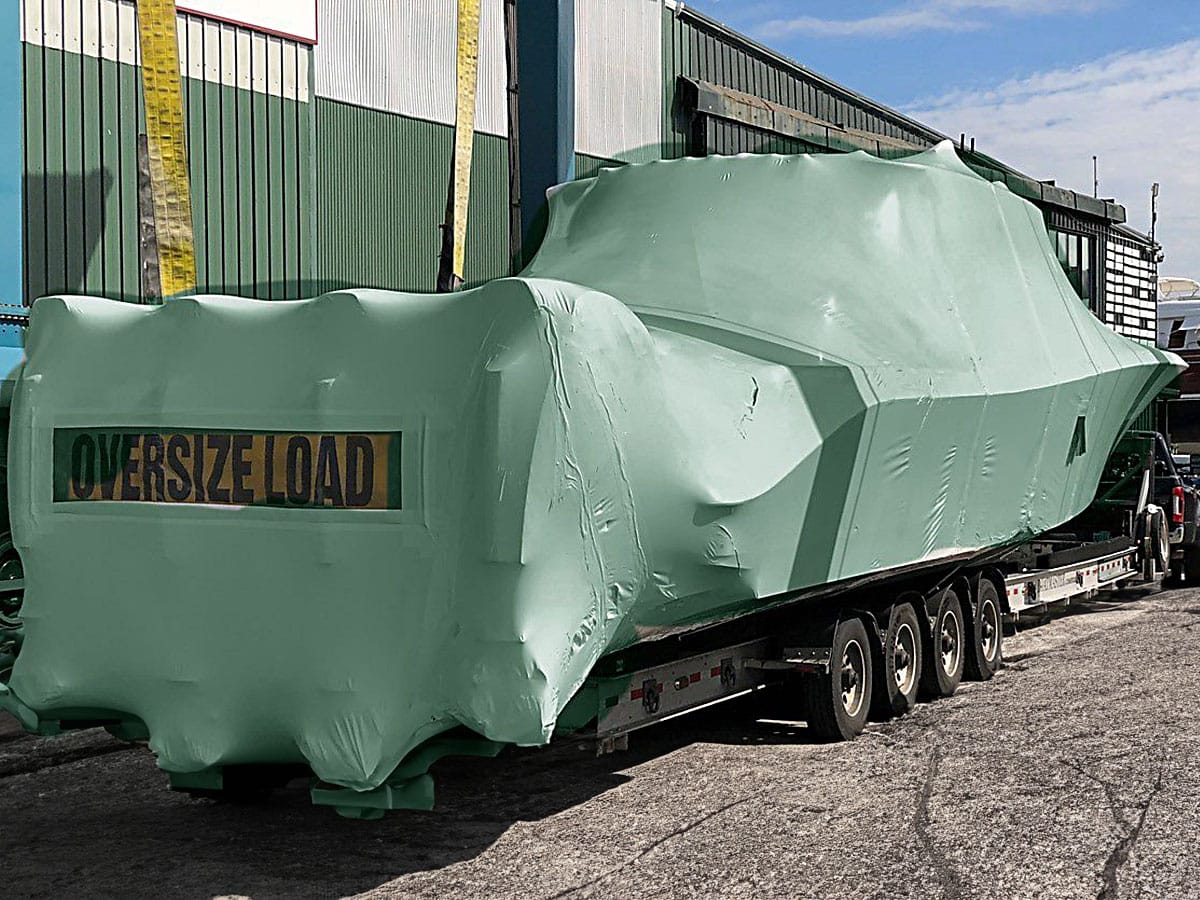 bioaqualife: A large boat, covered in green protective wrapping with the label "OVERSIZE LOAD," is secured on a trailer. The trailer is on a road near an industrial building. The sky is partly cloudy, and there are some industrial elements visible in the background.
