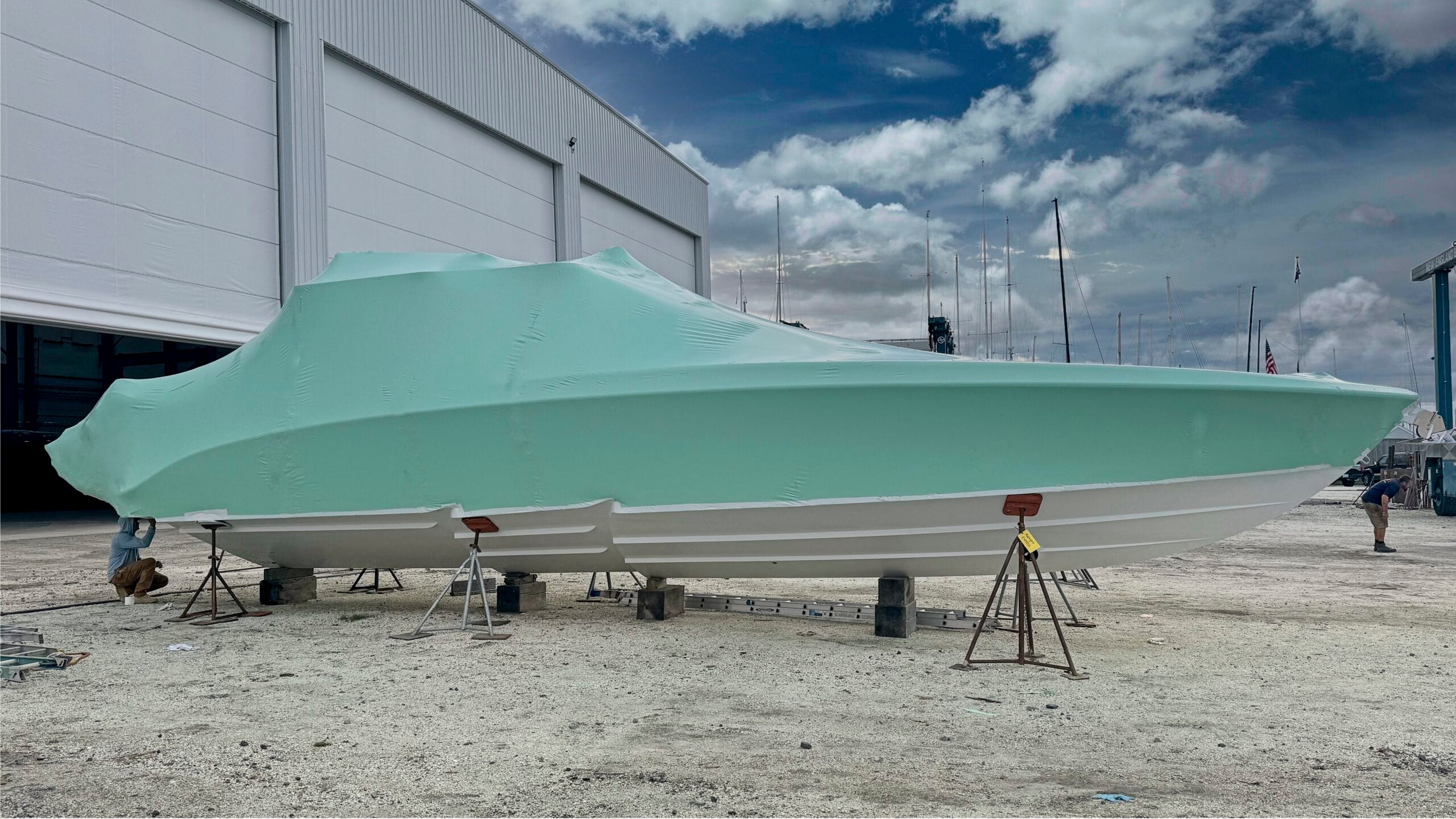 bioaqualife: A large boat covered in a light green tarp is propped up on stands in a boatyard. The boatyard is adjacent to a large white building with open doors, and several other masts and structures can be seen in the background under a partly cloudy sky.
