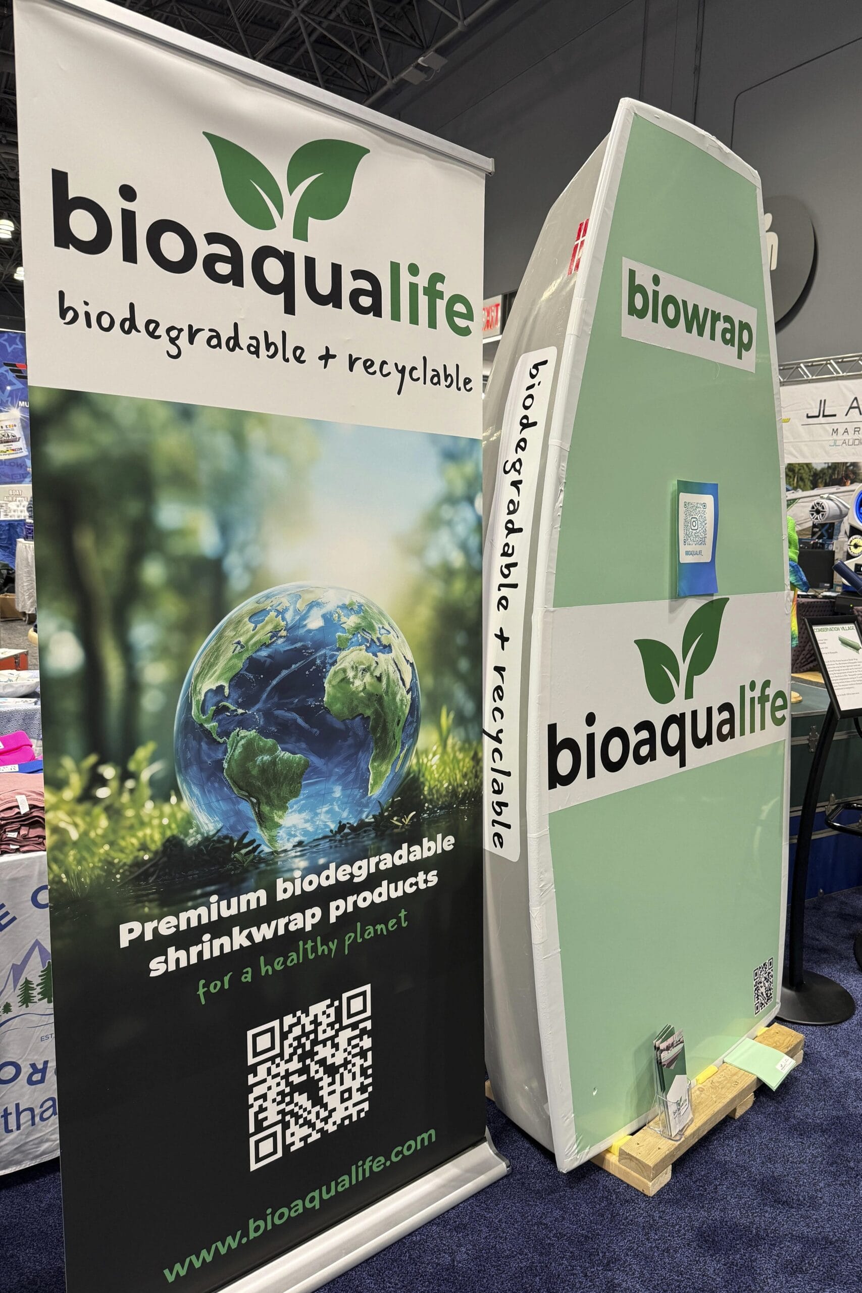 bioaqualife: A trade show booth for bioaqualife featuring a green and white surfboard-shaped display and a banner. The banner promotes biodegradable and recyclable shrink wrap products, with an image of Earth, a QR code, and the Bioaqualife website URL.