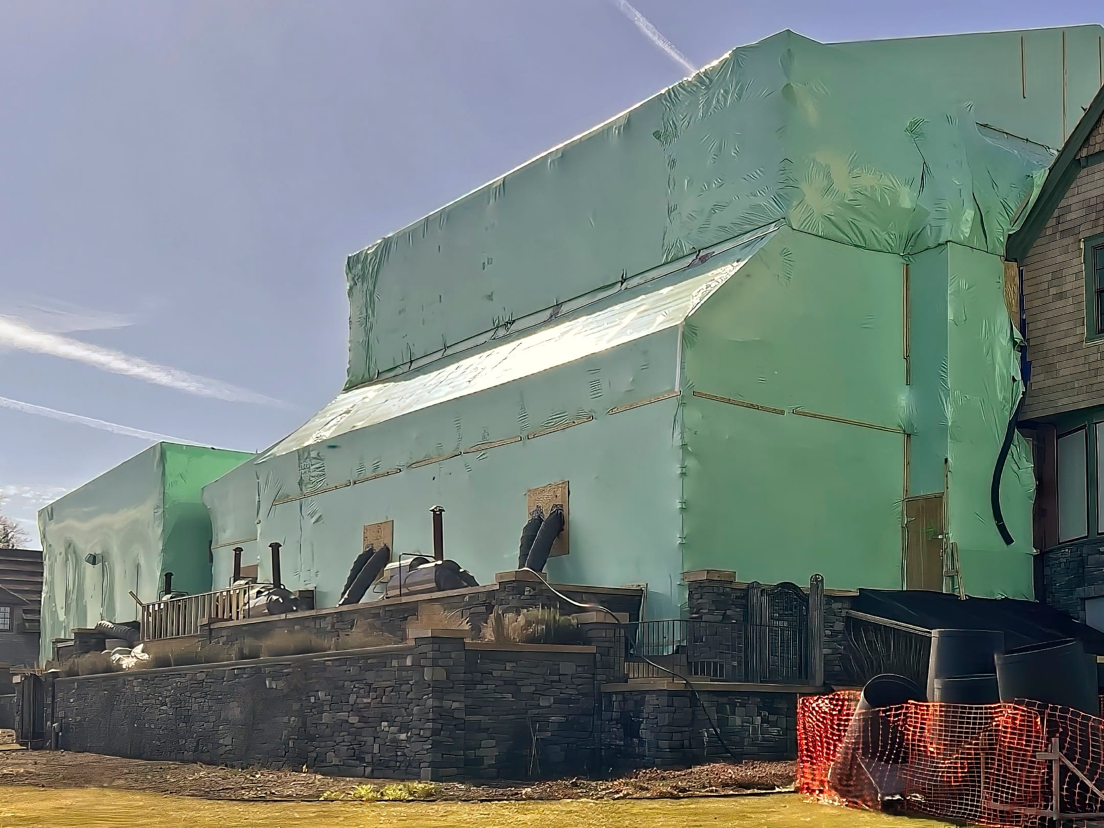 bioaqualife: A large building under construction is wrapped in green sheathing material. Stonework is visible at the base, and construction materials are scattered nearby. The sky is clear and blue with visible contrails.