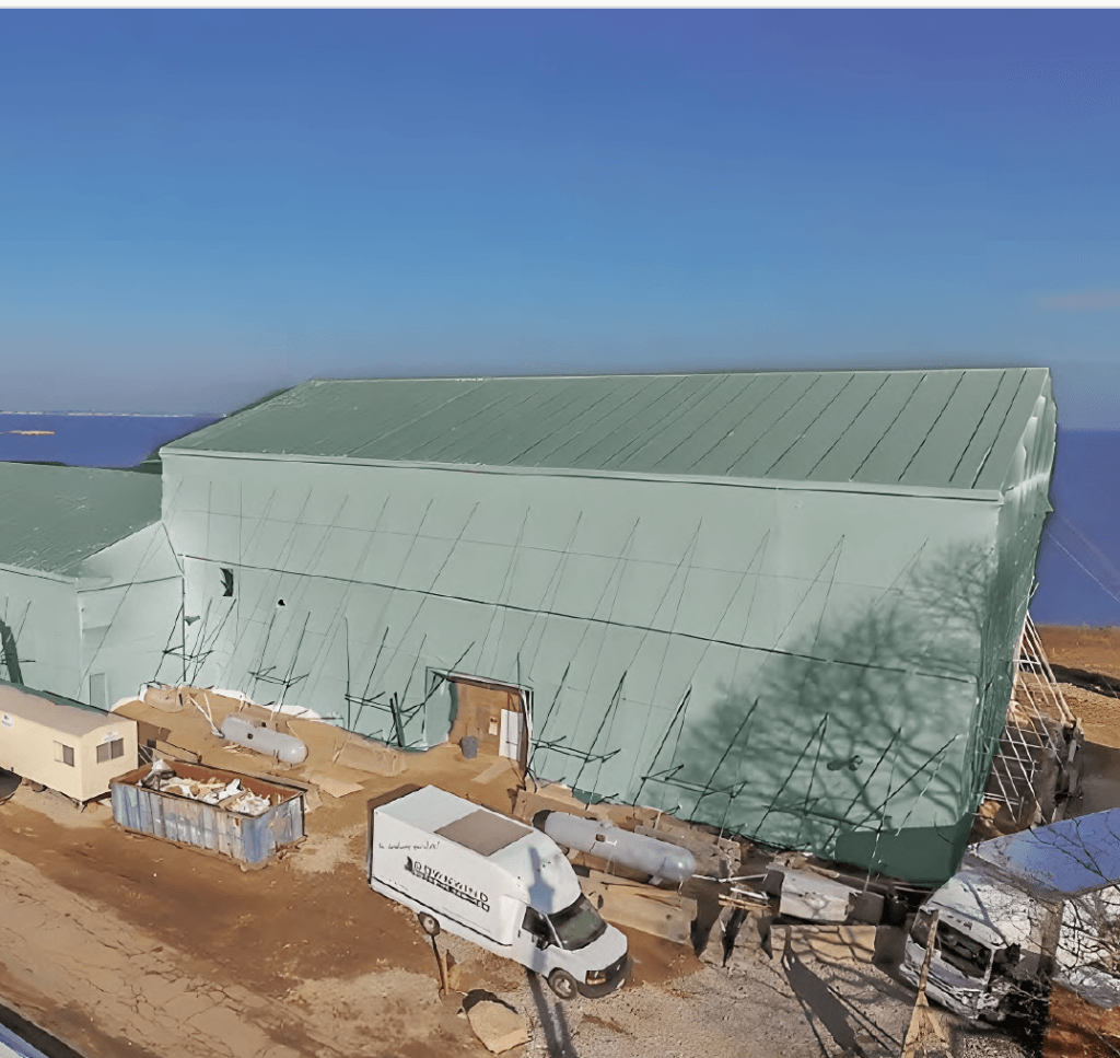 bioaqualife: A large building wrapped in green material, possibly undergoing construction or renovation. Several vehicles and containers are parked around it, under a clear blue sky.