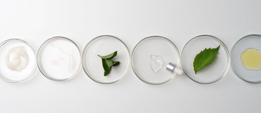 bioaqualife: A row of six circular glass dishes on a white surface containing different skincare ingredients: cream, liquid, two green leaves, a clear serum with a dropper, and another liquid.