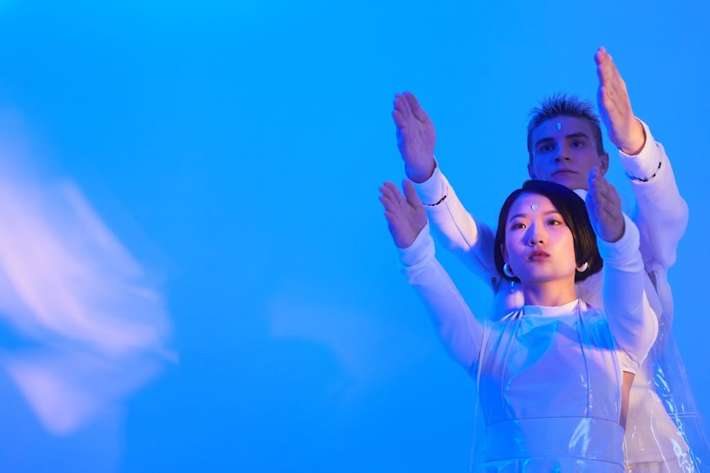 Young Man and Woman in Futuristic Clothing Standing in a Studio with Blue Lighting and Holding Their Arms Up