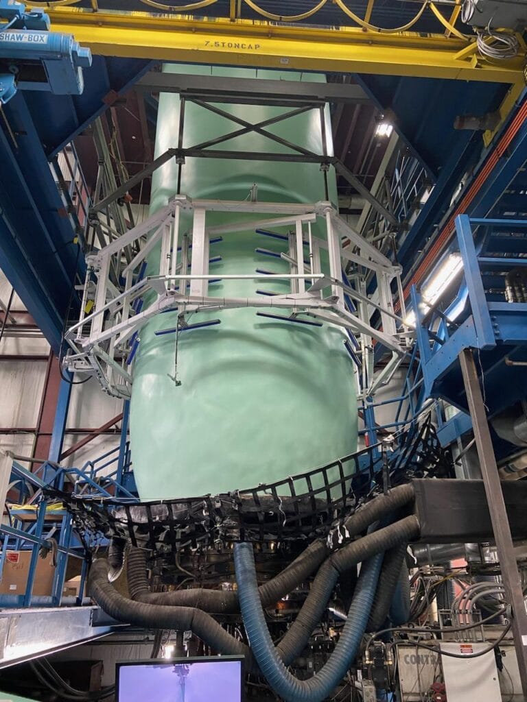 bioaqualife: A large industrial machine with a smooth, curved green surface surrounded by metal framing and cables. The structure is situated in a warehouse with blue beams and machinery components visible around it.