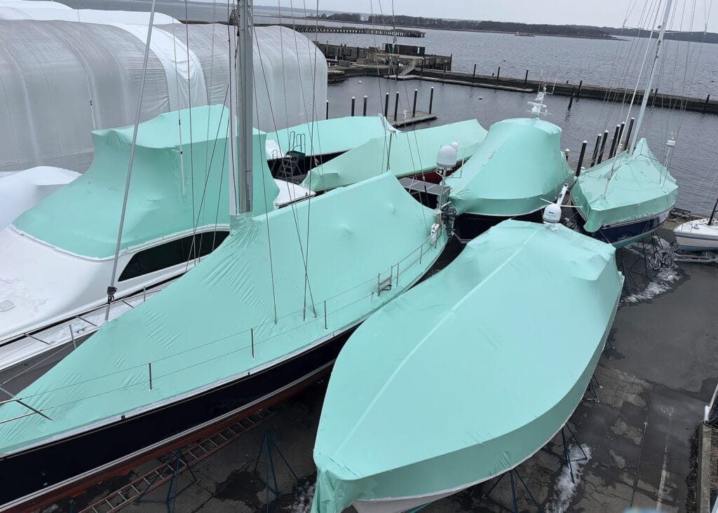 bioaqualife: Three boats covered in pale green tarps are docked at a marina. The surrounding area includes other covered boats and a view of the water with docks extending into it. The sky is overcast, creating a calm atmosphere.