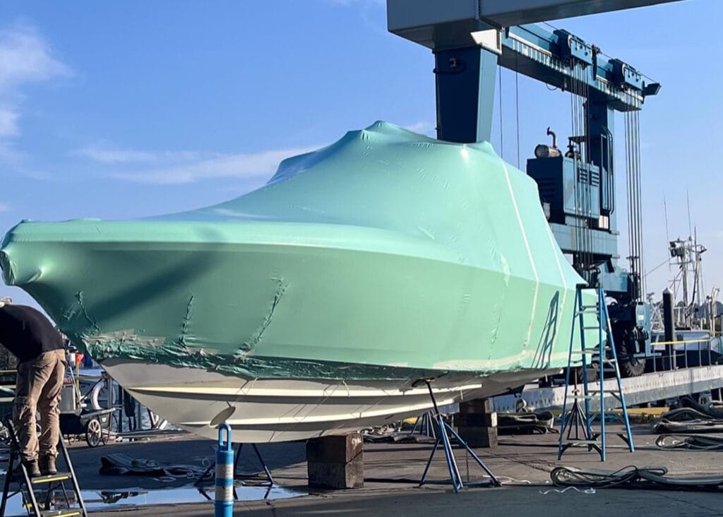 bioaqualife: A boat covered in green protective wrap is suspended in a shipyard by a large crane. Workers on ladders attend to the boat, with clear blue skies and other boats visible in the background.