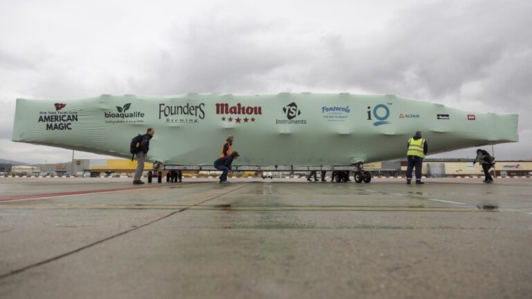 bioaqualife: Several people move a large, boat-shaped object covered by a green tarp printed with various sponsor logos across an airport tarmac on a cloudy day.