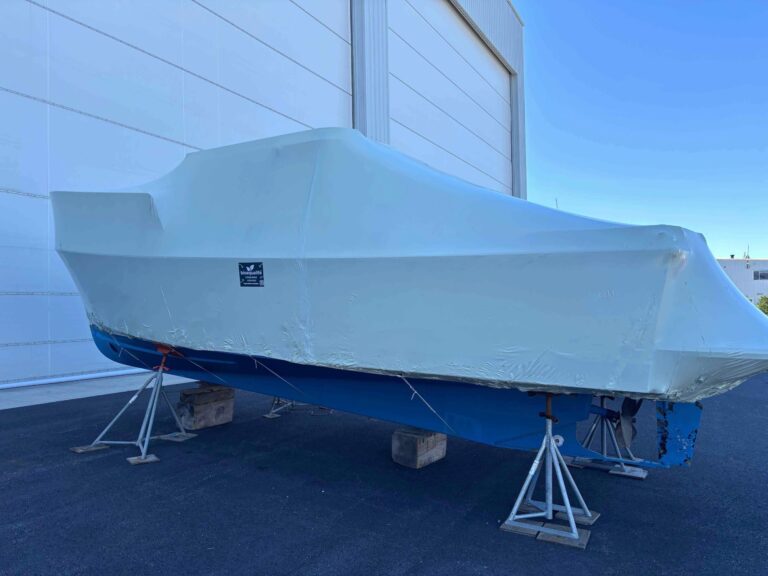 bioaqualife: A white and blue boat covered in protective shrink wrap is supported by metal stands and blocks beside a large building under a clear blue sky.