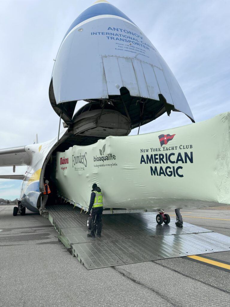 bioaqualife: A large cargo plane with its nose lifted open is being unloaded. Workers are guiding a wrapped yacht hull labeled &ldquo;American Magic, New York Yacht Club&rdquo; out of the aircraft onto a ramp.