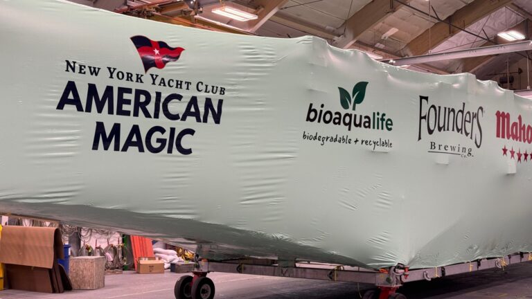 bioaqualife: A large boat covered in protective white wrap displays logos for the New York Yacht Club American Magic, bioaqualife, Founders Brewing, and Mahou inside an indoor facility.