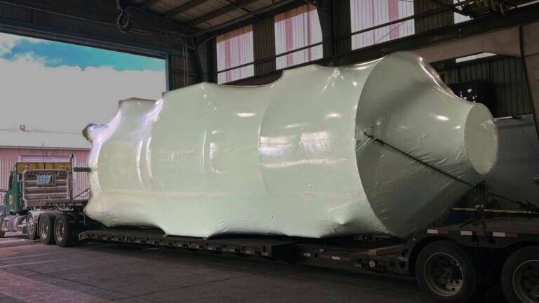 bioaqualife: A large, irregularly shaped industrial object wrapped in shiny white protective material is loaded onto a flatbed trailer inside a warehouse with a partially open roof.
