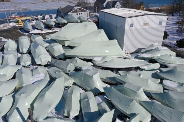 bioaqualife: Aerial view of numerous boats covered in white protective tarps, arranged closely together beside a white building on a snowy lot near a body of water.