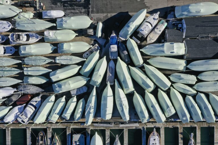 bioaqualife: Aerial view of numerous white boats arranged in rows on land near a dock, forming a fan-like pattern. Several boats are in water at the bottom of the image, while others are stored on the lot above.