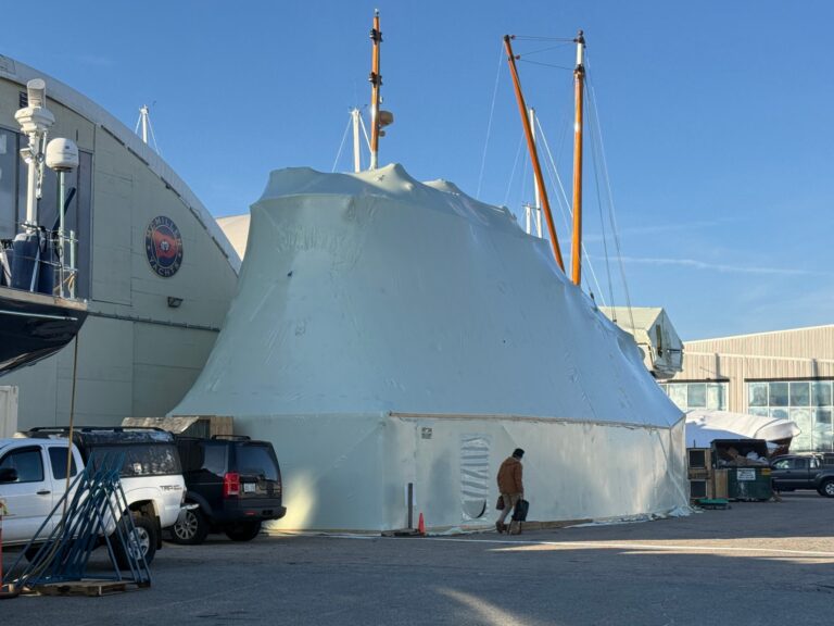 bioaqualife: A large boat covered in white shrink wrap sits outside a warehouse, with masts sticking out from the top. A person walks nearby, and several vehicles are parked along the building under a clear blue sky.