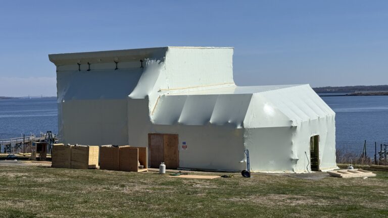 bioaqualife: A large, oddly shaped building is covered in pale insulation material near a body of water; wooden crates and construction materials are scattered on the grass around it under a clear blue sky.
