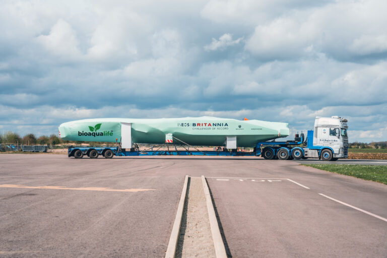 bioaqualife: A large white truck is transporting a long, green, covered racing yacht labeled "INEOS Britannia" and "bioaqualife" on a flatbed trailer, parked on an empty paved lot under a cloudy sky.