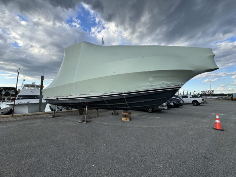 bioaqualife: A large boat covered in a protective wrap is propped up on stands in a marina parking lot, with a partly cloudy sky above and a traffic cone nearby.