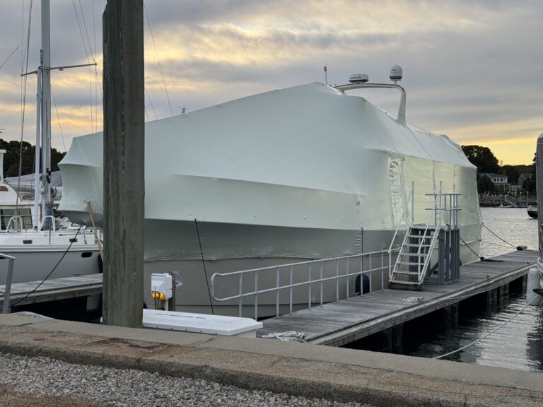 bioaqualife: A large boat covered in white shrink wrap is docked at a marina. There is a metal gangway attached to the boat and cloudy skies overhead. Other boats and houses are visible in the background.