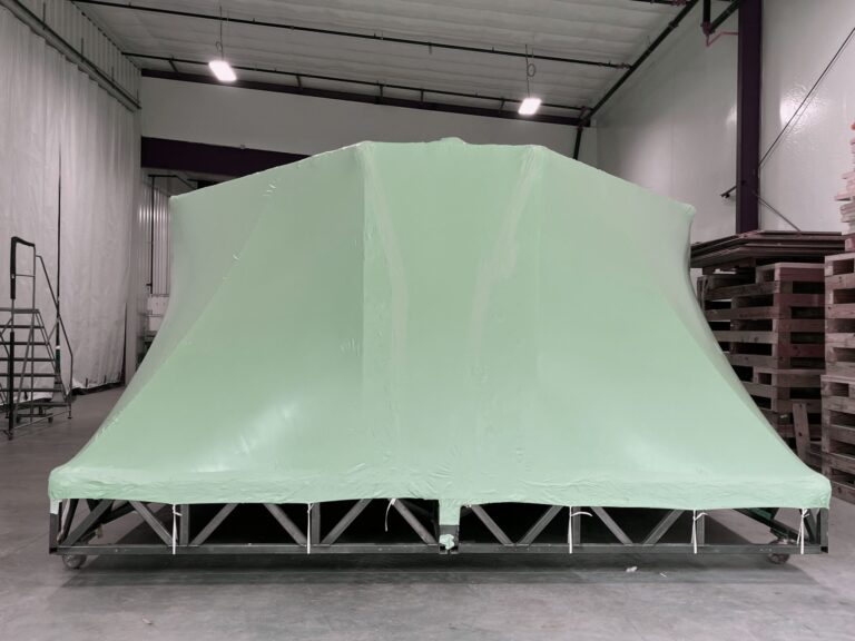 bioaqualife: A large, angular object is covered with smooth, green plastic sheeting in a spacious warehouse. The object rests on a metal frame, and stacks of wooden pallets are visible to the right.